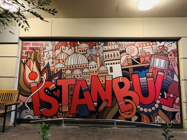A vibrant mural features the word 'Istanbul' in bold, red letters, surrounded by artistic depictions of iconic city landmarks and elements including domes, towers, and traditional symbols. The mural is colorful and displays a creative urban vibe. There is a wooden bench to the left of the mural and some leafy branches hanging down from the upper left corner.