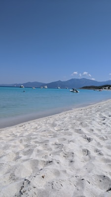 A serene beach scene with soft white sand and clear turquoise waters stretches into the distance. Several small boats and yachts are gently floating on the water. In the background, a range of mountains rises under a cloudless blue sky.