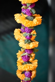 A colorful garland featuring alternating sections of bright orange marigold blossoms and purple globe amaranth flowers, set against a blurred dark green background. The garland is composed of tightly clustered flowers, creating a vibrant and visually striking pattern.