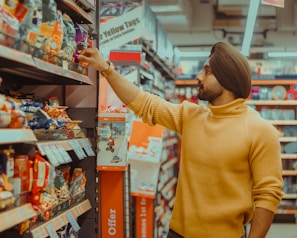 A person wearing a mustard colored sweater and a turban is reaching for an item on the shelf in a grocery store aisle. The shelves are stocked with various packaged snacks in colorful bags, and signage with promotional offers is visible.