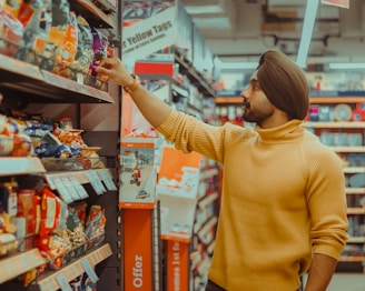 A person wearing a mustard colored sweater and a turban is reaching for an item on the shelf in a grocery store aisle. The shelves are stocked with various packaged snacks in colorful bags, and signage with promotional offers is visible.