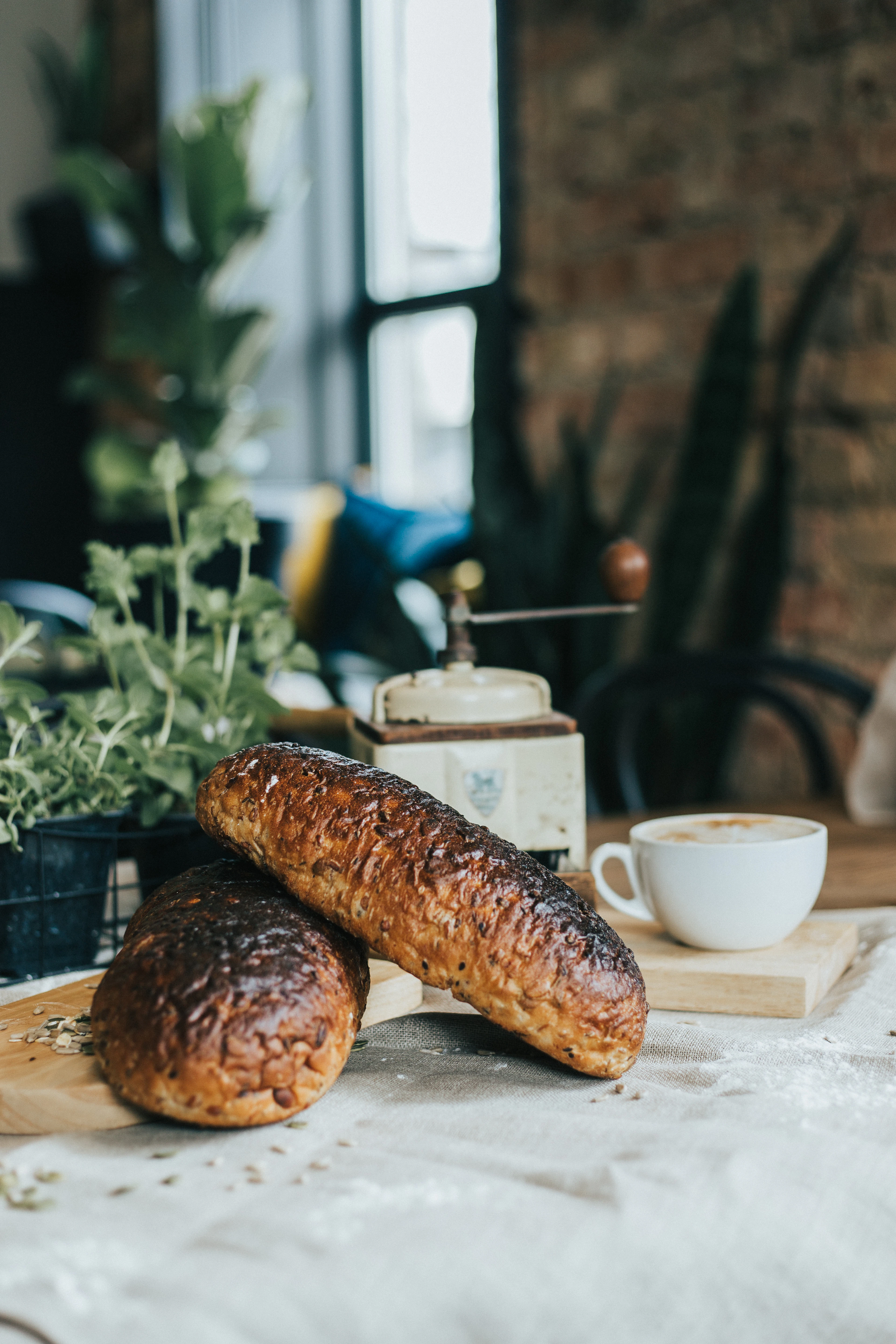 Two dark, crusty Latvian rye loaves resting on a wooden board beside a cup of coffee and an old hand grinder, lit by a window in a brick-walled room.