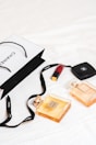 An arrangement of luxury items is laid out on a white surface, featuring two Chanel perfume bottles and a Chanel shopping bag. A red lipstick and a compact case are also present. The scene conveys a sense of elegance and sophistication with its minimalist composition.