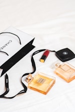 An arrangement of luxury items is laid out on a white surface, featuring two Chanel perfume bottles and a Chanel shopping bag. A red lipstick and a compact case are also present. The scene conveys a sense of elegance and sophistication with its minimalist composition.