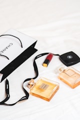 An arrangement of luxury items is laid out on a white surface, featuring two Chanel perfume bottles and a Chanel shopping bag. A red lipstick and a compact case are also present. The scene conveys a sense of elegance and sophistication with its minimalist composition.