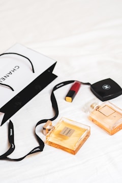 An arrangement of luxury items is laid out on a white surface, featuring two Chanel perfume bottles and a Chanel shopping bag. A red lipstick and a compact case are also present. The scene conveys a sense of elegance and sophistication with its minimalist composition.