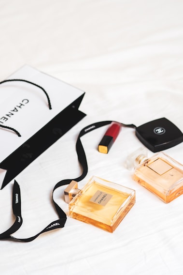 An arrangement of luxury items is laid out on a white surface, featuring two Chanel perfume bottles and a Chanel shopping bag. A red lipstick and a compact case are also present. The scene conveys a sense of elegance and sophistication with its minimalist composition.