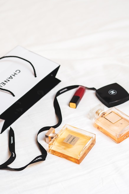 An arrangement of luxury items is laid out on a white surface, featuring two Chanel perfume bottles and a Chanel shopping bag. A red lipstick and a compact case are also present. The scene conveys a sense of elegance and sophistication with its minimalist composition.