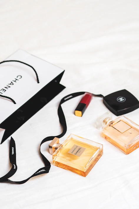 An arrangement of luxury items is laid out on a white surface, featuring two Chanel perfume bottles and a Chanel shopping bag. A red lipstick and a compact case are also present. The scene conveys a sense of elegance and sophistication with its minimalist composition.