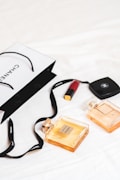 An arrangement of luxury items is laid out on a white surface, featuring two Chanel perfume bottles and a Chanel shopping bag. A red lipstick and a compact case are also present. The scene conveys a sense of elegance and sophistication with its minimalist composition.