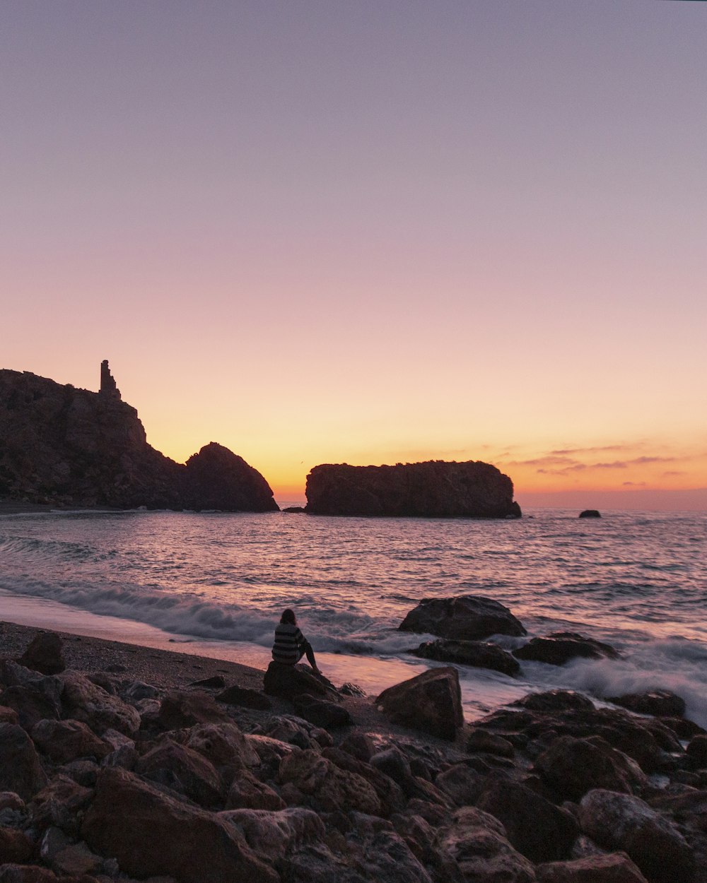 pessoa sentada em formação rochosa perto do mar durante o pôr do sol