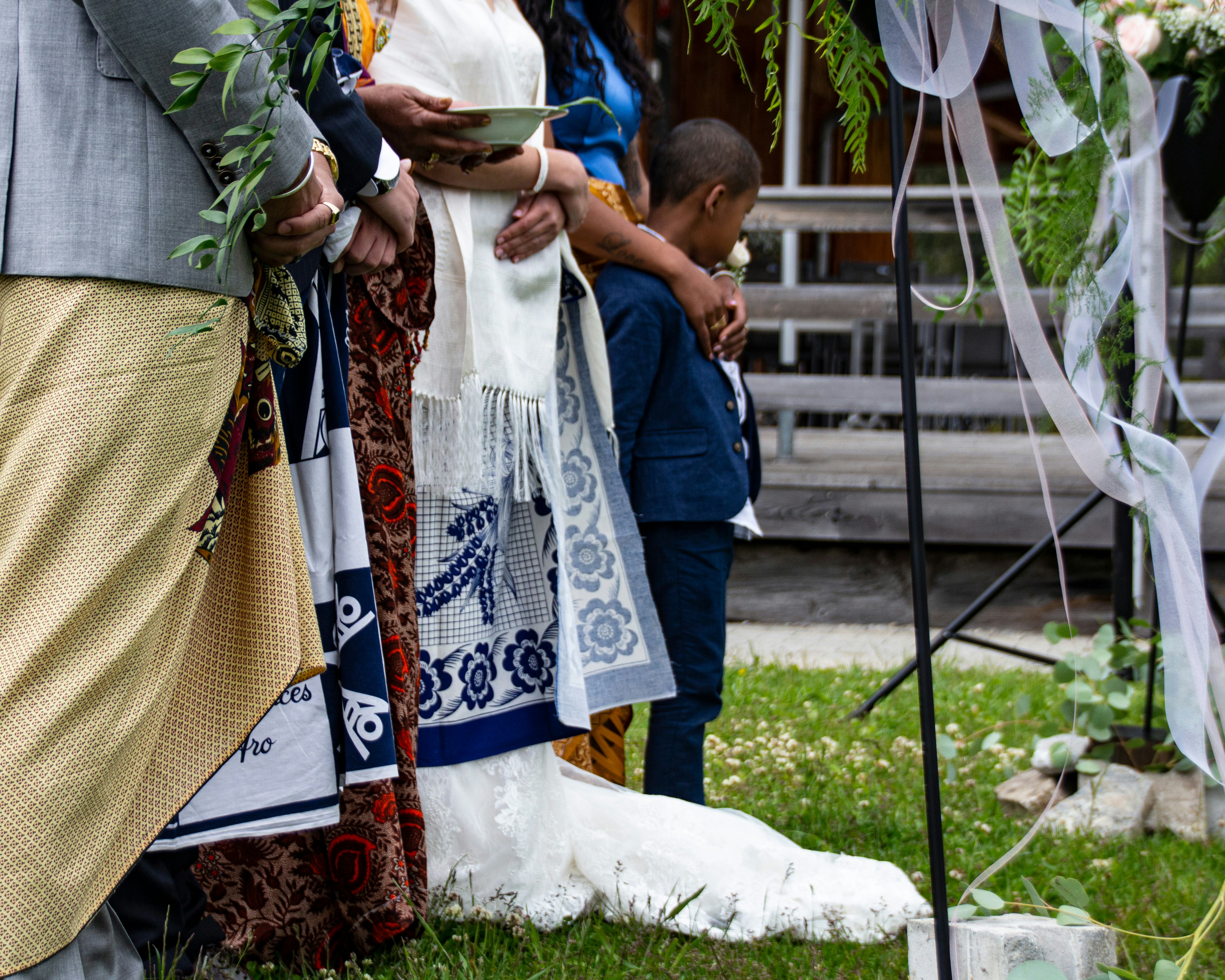 A group of people stand together, adorned in colorful traditional attire, likely participating in a formal or ceremonial event. One person holds a leafy branch, while another cradles a young boy, indicating a sense of community or family. The setting includes decorative elements such as draped ribbons and greenery, suggesting a festive or celebratory occasion.
