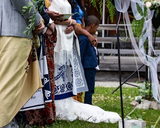 A group of people stand together, adorned in colorful traditional attire, likely participating in a formal or ceremonial event. One person holds a leafy branch, while another cradles a young boy, indicating a sense of community or family. The setting includes decorative elements such as draped ribbons and greenery, suggesting a festive or celebratory occasion.
