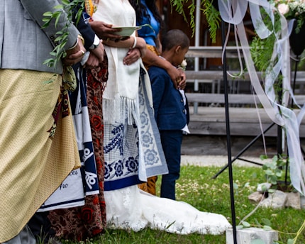 A group of people stand together, adorned in colorful traditional attire, likely participating in a formal or ceremonial event. One person holds a leafy branch, while another cradles a young boy, indicating a sense of community or family. The setting includes decorative elements such as draped ribbons and greenery, suggesting a festive or celebratory occasion.
