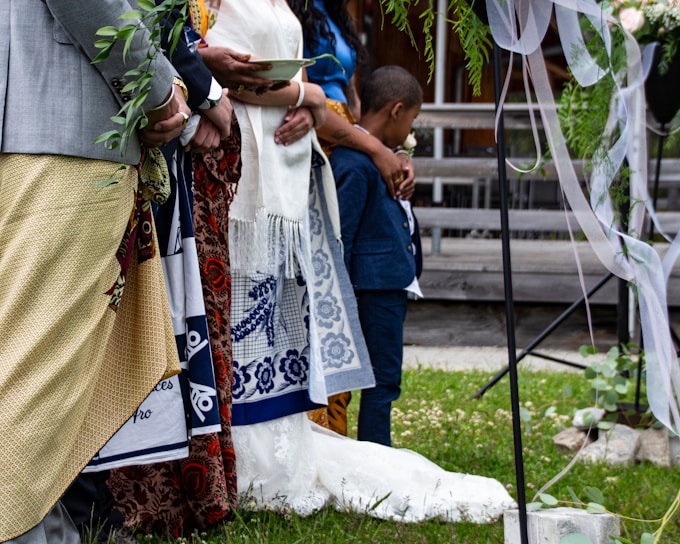 A group of people stand together, adorned in colorful traditional attire, likely participating in a formal or ceremonial event. One person holds a leafy branch, while another cradles a young boy, indicating a sense of community or family. The setting includes decorative elements such as draped ribbons and greenery, suggesting a festive or celebratory occasion.
