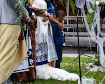 A group of people stand together, adorned in colorful traditional attire, likely participating in a formal or ceremonial event. One person holds a leafy branch, while another cradles a young boy, indicating a sense of community or family. The setting includes decorative elements such as draped ribbons and greenery, suggesting a festive or celebratory occasion.