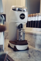 A modern white coffee grinder with the brand name 'Niche' displayed on it sits atop a marble countertop. Behind the grinder, there is a row of clear glass jars filled with coffee beans. The grinder has wooden accents, and a metal container is placed underneath its spout.