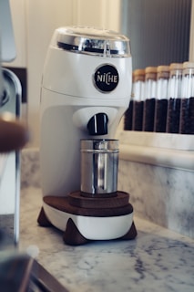 A modern white coffee grinder with the brand name 'Niche' displayed on it sits atop a marble countertop. Behind the grinder, there is a row of clear glass jars filled with coffee beans. The grinder has wooden accents, and a metal container is placed underneath its spout.