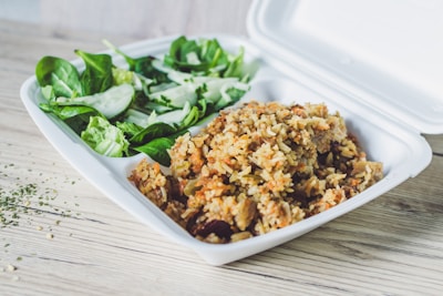 A takeout container filled with a serving of mixed rice on one side and a green salad on the other. The salad includes spinach leaves and sliced cucumber, while the rice appears to be mixed with vegetables or meat. The container is placed on a light wooden surface, and a sprinkle of herbs is visible nearby.