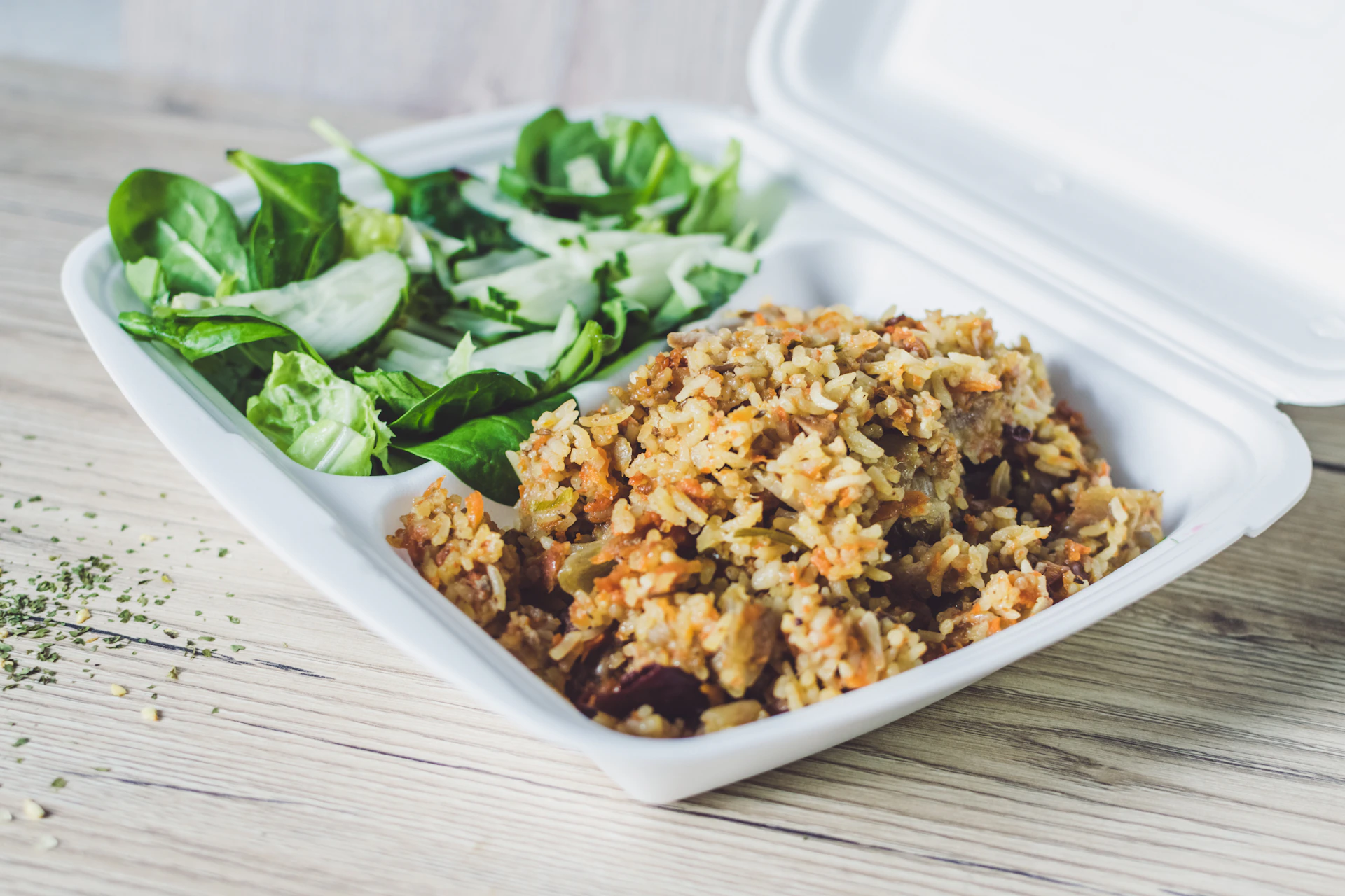A takeout container filled with a serving of mixed rice on one side and a green salad on the other. The salad includes spinach leaves and sliced cucumber, while the rice appears to be mixed with vegetables or meat. The container is placed on a light wooden surface, and a sprinkle of herbs is visible nearby.