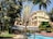 A hotel courtyard with a blend of natural and architectural elements, including a swimming pool in the foreground, surrounded by lush green trees and a yellow multi-story building in the background. There are wicker chairs and tables near the pool, under a canopy. Palm trees and other tropical plants add to the serene environment.