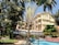 A hotel courtyard with a blend of natural and architectural elements, including a swimming pool in the foreground, surrounded by lush green trees and a yellow multi-story building in the background. There are wicker chairs and tables near the pool, under a canopy. Palm trees and other tropical plants add to the serene environment.