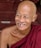 An elderly person with a bald head is smiling broadly. Wearing a maroon robe, they sit on red stone steps. Their expression is joyful and relaxed, showing deep wrinkles that add character to their smiling face.