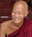 An elderly person with a bald head is smiling broadly. Wearing a maroon robe, they sit on red stone steps. Their expression is joyful and relaxed, showing deep wrinkles that add character to their smiling face.