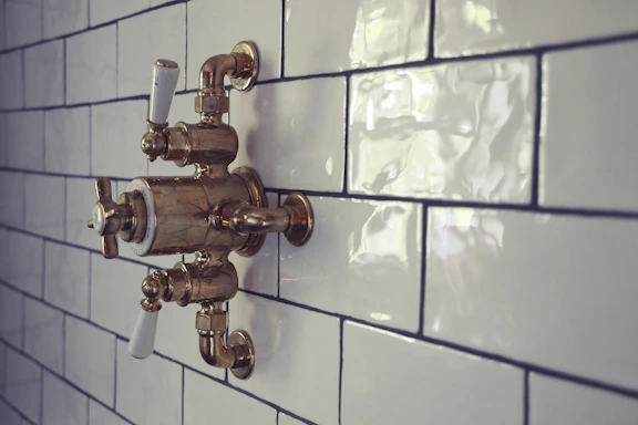 A vintage-style bronze shower valve with white handles is mounted on a glossy white subway-tiled wall. The tiles are rectangular and feature a subtle sheen, accentuating the classic and polished aesthetic of the bathroom fixture.