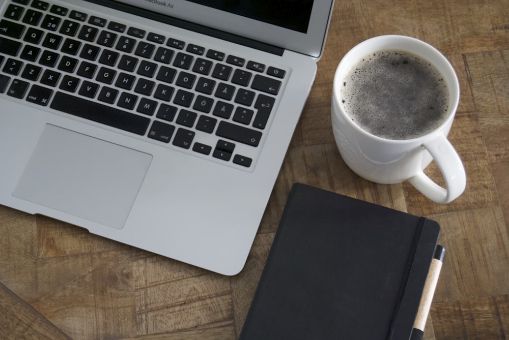 macbook pro ao lado da caneca de cerâmica branca na mesa de madeira marrom