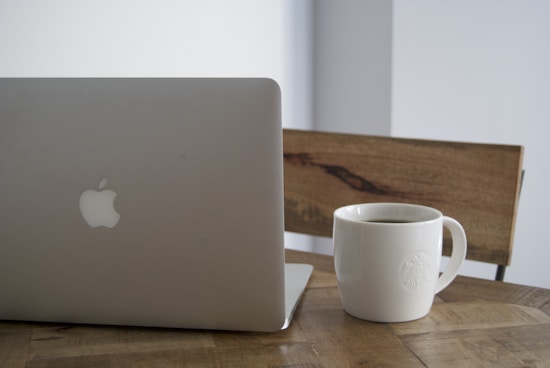 A sleek laptop with a visible logo is placed on a wooden table next to a white coffee mug filled with a dark liquid. The setting appears to be a minimalistic and modern workspace with a wooden chair in the background.