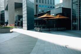 A modern urban setting featuring tall buildings and a shaded outdoor seating area with bright orange umbrellas. The architecture displays a mix of concrete and glass elements with geometric patterns. Sunlight creates contrasting shadows across the tiled pavement. A small garden bed filled with red flowers adds a touch of nature to the scene.