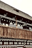 A traditional wooden building is adorned with intricate white geometric patterns on its facade. The structure features a wooden balcony with vertical slats and a steeply pitched wooden roof. The patterns include spirals, chevrons, and heart shapes.