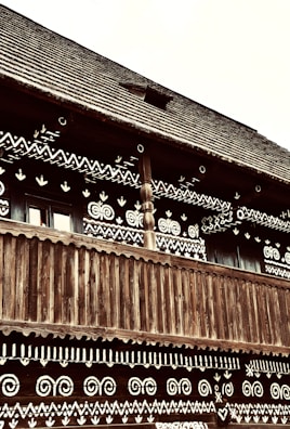 A traditional wooden building is adorned with intricate white geometric patterns on its facade. The structure features a wooden balcony with vertical slats and a steeply pitched wooden roof. The patterns include spirals, chevrons, and heart shapes.