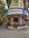 A corner building features a restaurant with a blue and white sign reading 'Koolar & Co. Restaurant & Stores' in English and Hindi. The structure has an old, rustic appearance with a round facade and a small patio area where people are sitting. Surrounding the building are trees and other structures, with a few people walking along the street.