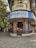 A corner building features a restaurant with a blue and white sign reading 'Koolar & Co. Restaurant & Stores' in English and Hindi. The structure has an old, rustic appearance with a round facade and a small patio area where people are sitting. Surrounding the building are trees and other structures, with a few people walking along the street.