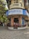 A corner building features a restaurant with a blue and white sign reading 'Koolar & Co. Restaurant & Stores' in English and Hindi. The structure has an old, rustic appearance with a round facade and a small patio area where people are sitting. Surrounding the building are trees and other structures, with a few people walking along the street.