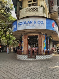 A corner building features a restaurant with a blue and white sign reading 'Koolar & Co. Restaurant & Stores' in English and Hindi. The structure has an old, rustic appearance with a round facade and a small patio area where people are sitting. Surrounding the building are trees and other structures, with a few people walking along the street.
