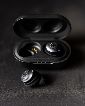A pair of black wireless earbuds are placed in an open charging case. One earbud is positioned outside the case on a dark fabric surface. The earbuds have a sleek design with small blue indicators.