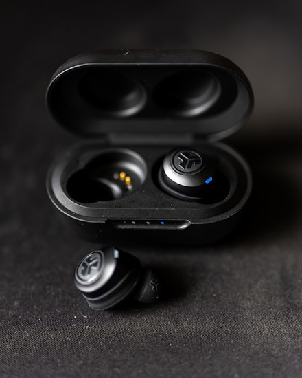 A pair of black wireless earbuds are placed in an open charging case. One earbud is positioned outside the case on a dark fabric surface. The earbuds have a sleek design with small blue indicators.
