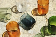 Various colored glass cups are arranged on a textured surface. The cups cast distinct shadows due to the sunlight coming from an angle. The glasses include shades of orange, green, clear, and blue. Each cup has a unique texture or pattern on its surface.