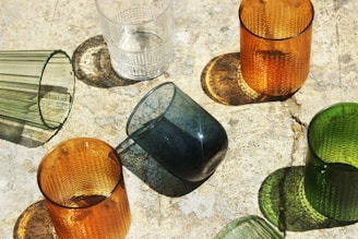 Various colored glass cups are arranged on a textured surface. The cups cast distinct shadows due to the sunlight coming from an angle. The glasses include shades of orange, green, clear, and blue. Each cup has a unique texture or pattern on its surface.