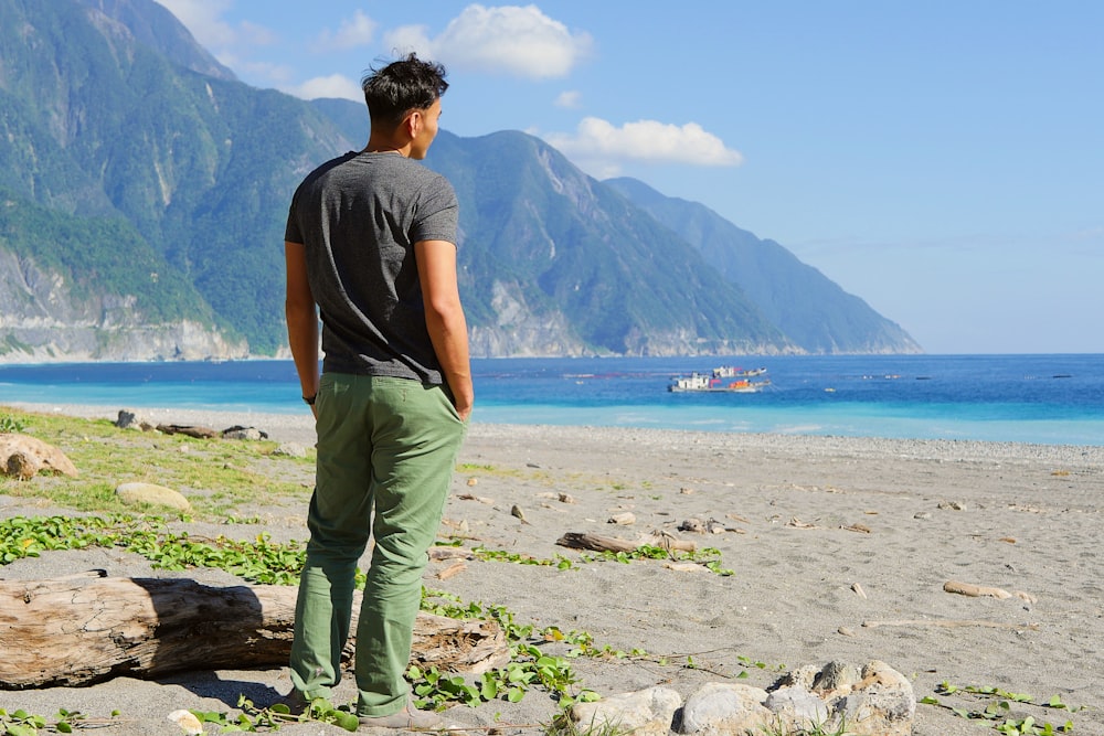 man in gray t-shirt and green pants standing on gray rock near body of water