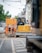 A road construction scene with several workers dressed in bright orange vests and hard hats. They are standing near a large yellow construction vehicle with buildings lining the street in the background. Overhead, there's a network of cables, possibly for trams, and there is visible construction material on the ground.