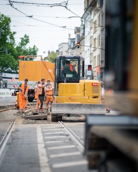 A road construction scene with several workers dressed in bright orange vests and hard hats. They are standing near a large yellow construction vehicle with buildings lining the street in the background. Overhead, there's a network of cables, possibly for trams, and there is visible construction material on the ground.