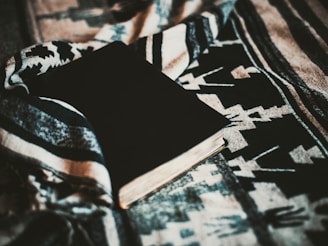 A closed book with a dark cover lies on a fabric with an intricate pattern that features geometric shapes. The fabric appears textured and tactile, with a mix of earthy and neutral tones that enhance its visual appeal.