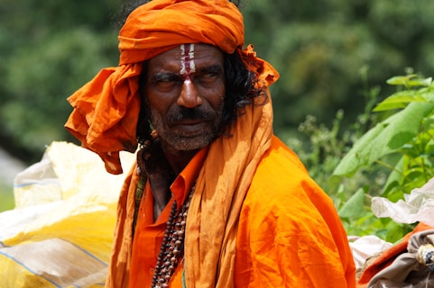 A man wearing a bright orange robe and turban with a traditional bead necklace, standing outdoors. He has facial markings and appears contemplative, surrounded by greenery.