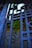 Intricately designed wrought iron bars with a geometric pattern are set against a backdrop where green foliage is visible through the gaps. The metal has a slightly weathered texture, suggesting age or exposure to elements.