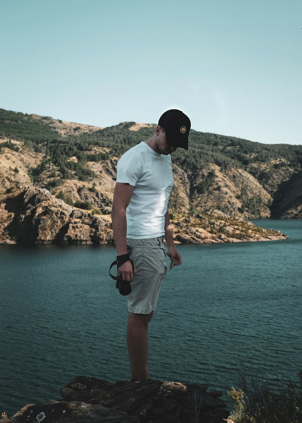man in white t-shirt and brown shorts standing on rock formation near body of water