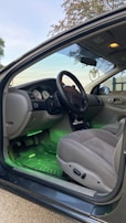 The image displays the interior of a car from the driver's side with the door open. The dashboard features a steering wheel, speedometer, and instrument panels. The interior has gray upholstery and a rubber floor mat with green ambient lighting underneath the dashboard.